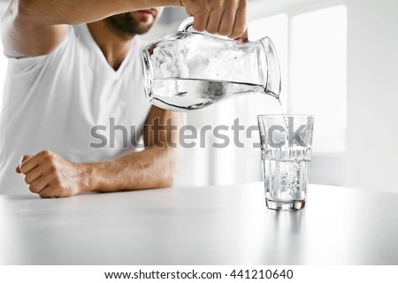 Drink Water. Close Up Of Handsome Young Man Pouring Fresh Pure Water From Pitcher Into A Glass In Morning In Kitchen. Beautiful Athletic Male Model Feeling Thirsty. Healthy Nutrition And Hydration
