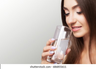Drink water. Happy young beautiful woman drinking water. A smiling female model holding a transparent glass in his hand. Focus on your hand. On a gray background. - Shutterstock ID 686855611
