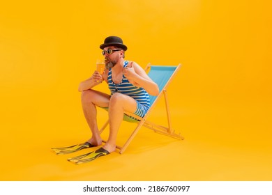 Drinking Beer. Studio Shoot Of Cool Funny Beachgoer, Man In Vintage Striped Swimsuit And Bowler Hat Sitting In Sunbed Isolated On Yellow Background. Leisure Activity, Vacation, Summer, Sales