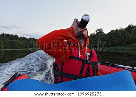 Drinking captain man in lifejacket with a bottle of beer in his hand on the stern of a fast gliding red PVC inflatable boat floats on the river at summer evening, the danger of drunken navigation
