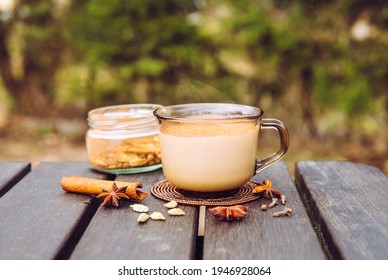 Drinking Chai Latte At Picnic Outdoors Concept. Cup Of Warm Chai Latte On Picnic Table In Campsite, Jar With Spice Mixture On Background.