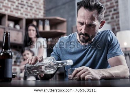 Drinking father. Serious unhappy sad man sitting at the table and pouring vodka into his glass while having alcohol addiction