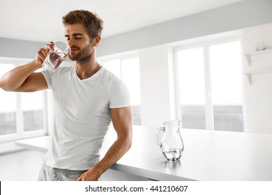 Drinking Water. Closeup Portrait Of Handsome Young Man With Sexy Fit Body Drinking Fresh Water From Glass In Morning. Thirsty Fitness Male Model Enjoying Refreshing Drink Indoors. Healthy Nutrition
