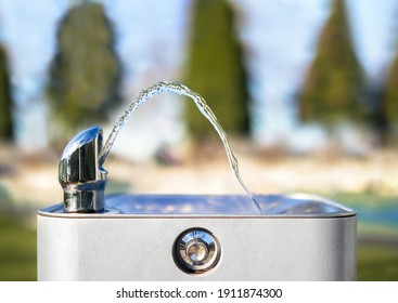 Drinking Water Fountain In Park On A Sunny Day, No Person. Close Up Of Flowing Water From The Tab In An Arch. Bright Defocused Park And Tree Background. Selective Focus. 