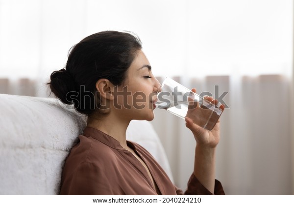 Drinking water. Side view young indian woman enjoy\
pure fresh cool mineral water at morning. Profile shot of thirsty\
millennial mixed race lady hold glass swallow crystal still aqua\
with closed eyes