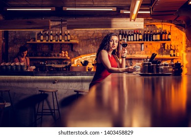 Drinking Wine Alone. Stylish Curly Woman Wearing Red Dress Sitting At The Bar And Drinking Wine Alone