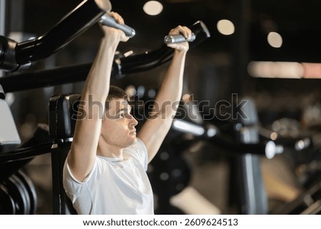 Driven young man training on shoulder press machine in modern well-equipped gym, focused on working deltoids, building upper body strength and staying committed to fit lifestyle