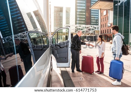 Driver of a airport shuttle minivan, greeting his passengers with their luggage on the sidewalk of a modern city business district