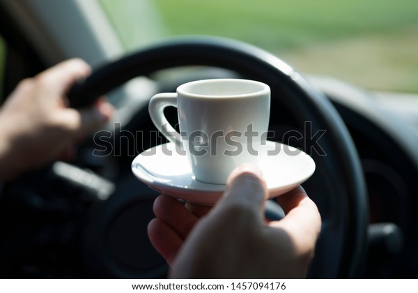 The driver behind the wheel of the car. One\
driver\'s hand holds the steering wheel, the other hand holds a\
white coffee Cup. Selective focus. Is defocused. Concept: coffee\
with you on a road trip.