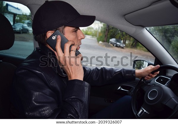 A driver in a black\
cap and a leather jacket is talking on the phone in a car driving\
on an autumn evening