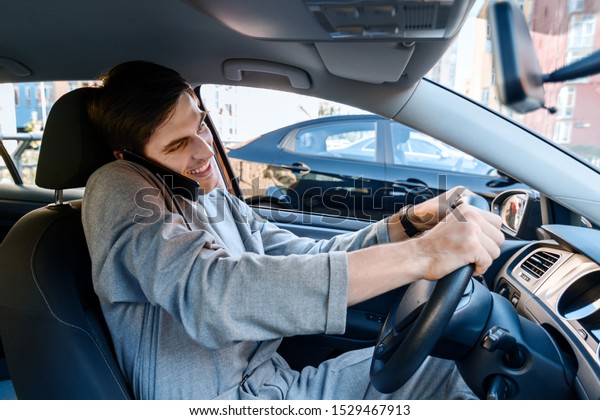 Driver businessman is talking on smartphone behind\
steering wheel of car. Young man is not attentively driving\
automobile. Guy is breaking rules of road. Concept of fast rhythm\
in modern city.