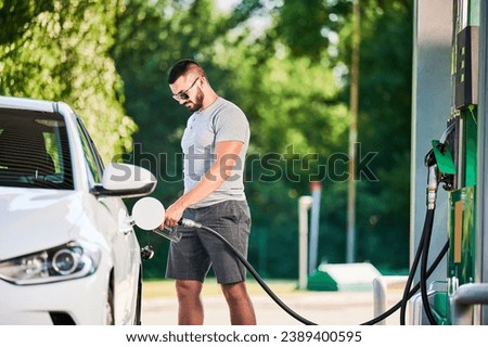 Driver with gasoline pump refilling car gas tank. Confident man refueling his luxury white auto. Man in casual clothes and sunglasses at modern gas station.