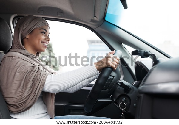 Driver goes to office in city, lesson and test\
drive. Smiling young pretty islamic african-american female in\
hijab in car on steering wheel, enjoying travel or trip in auto,\
side view, free space
