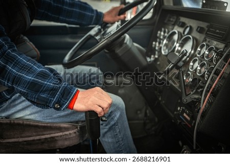 A driver grips the steering wheel while shifting gears inside a large truck's cabin. The environment is focused and bustling with activity.