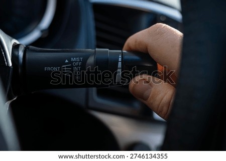 Driver hand adjusting windshield wiper control lever on steering column inside car interior, selecting mist and interval modes.