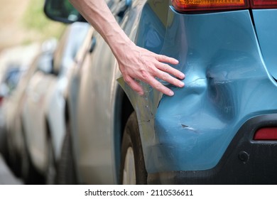 Driver Hand Examining Dented Car With Damaged Fender Parked On City Street Side. Road Safety And Vehicle Insurance Concept