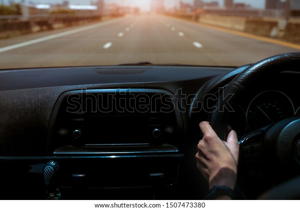 Driver hand holding steering wheel for control car.\
Inside view of car. Dashboard and windshield. Driving car on\
concrete road in the city. Perspective view car on highway. Driver\
hand wear smart band