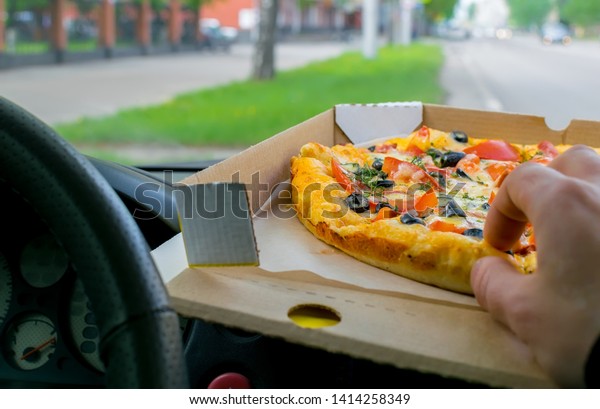 the driver hand takes a piece of pizza, the\
packaging of which lies on the windshield panel of the parked car\
on the background of the city\
roadway
