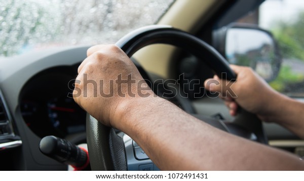 Driver\
holding steering wheel and drive on raining. He driving car careful\
after raining on mirror car. Hand of old human control car and\
looking transport on road in heavy weather\
