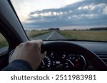 A driver holds the steering wheel, gazing at an open countryside road under a cloudy sky, showcasing a serene drive through rural landscapes.