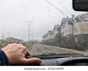 Driver holds steering wheel while driving in heavy rain through city streets with a wet windshield and visible traffic.