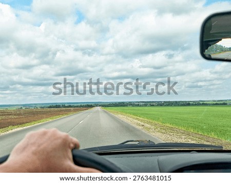 A driver holds the steering wheel while looking ahead at the road lined with fields on a cloudy but sunny day.