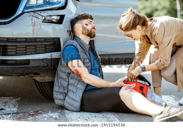 Driver hurrying with first aid kit to help\
injured man with bleeding wounds sitting near the car after the\
road accident