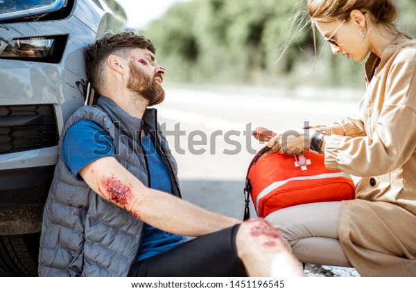 Driver hurrying with first aid kit to help\
injured man with bleeding wounds sitting near the car after the\
road accident