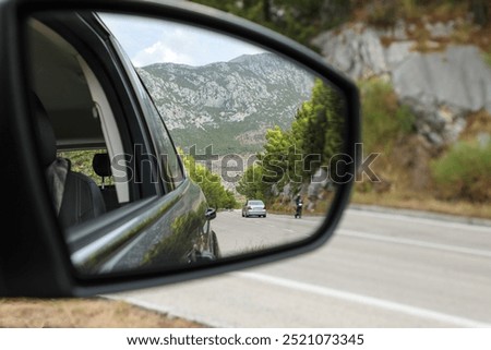 The driver looks into the side rearview mirror of the car and sees the road, high mountains and other road users.