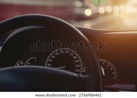 A driver looks at the speedometer while driving on a road in the evening. The sun emits a warm glow as cars pass by in the background, creating a sense of motion and normal activity. Copy ad text space