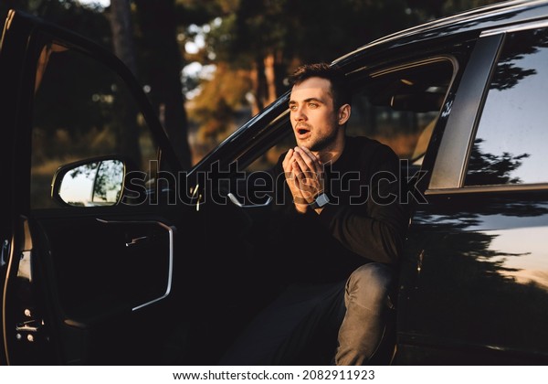 Driver man sitting in the black car on nature and
look shocked. Ordinary slender european male model with beard
holding hands on cheeks with opened mouth and shocked expression,
being shock and scare.