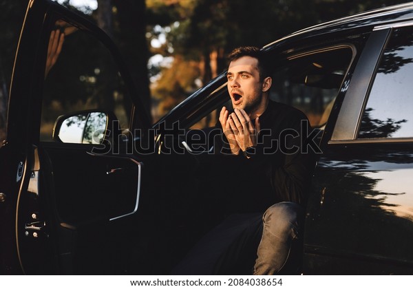 Driver man sitting in the black car on nature and\
look shocked. Ordinary slender european male model with beard\
holding hands on cheeks with opened mouth and shocked expression,\
being shock and scare.