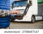 Driver managing logistics, fleet, and delivery scheduling with a laptop in front of a heavy goods vehicle
