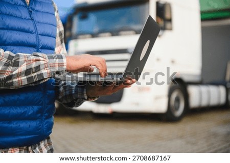 Driver managing logistics, fleet, and delivery scheduling with a laptop in front of a heavy goods vehicle