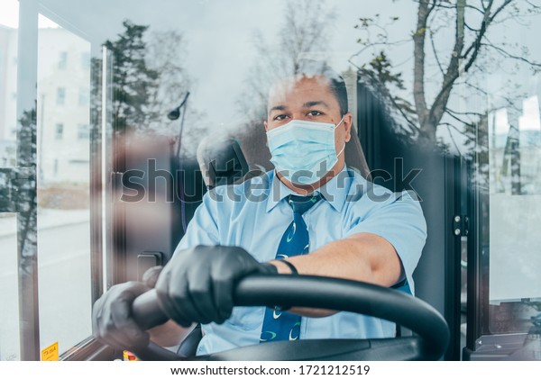 driver in mask looks at road while driving. young\
latin man bus driver in blue shirt has blue medical protection mask\
and black  gloves on hands.  bus driver wants to prevent  infection\
of covid 19.