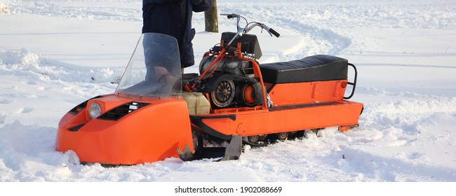 A Driver Near Orange Old Broken Russian Utility Single Ski Snowmobile Without Cover Close Up - Steering, Engine Handle And Fuel Tank On White Snow At Winter Day