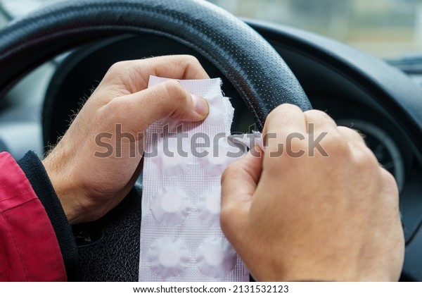 The driver opens a package of pills against a\
blurred background of the steering wheel in the car. The use of\
pharmacological drugs for medical purposes while driving. Selective\
focus, toned