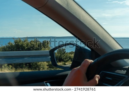 Driver perspective view from inside car driving along coastal road with blue sea and horizon on sunny summer day. Concept of road trip, travel freedom, vacation journey and scenic landscape. 