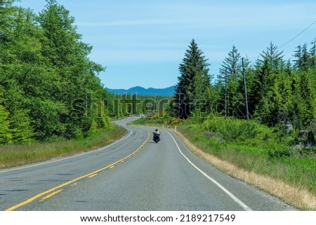 Driver perspective view of motorcycle in front with passenger (pillion) on otherwise empty curving two lane road in mountainous and forested USA landscape against a blue sky
