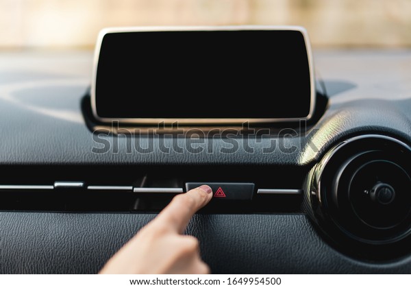 Driver pressing on hazard button on a car\
dashboard close up with empty infotainment screen (entertainment\
screen inside a car). Woman pushing on red hazard button on car\
dashboard with empty\
monitor.
