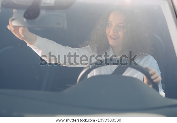 Driver in rear-view mirror. Attractive young woman\
in business wear looking in rear-view mirror and smiling while\
driving a car.