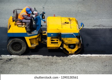 Driver of road roller leveling new layer of asphalt. Not large machine in yellow and blue colors. Road roller without the cabine.