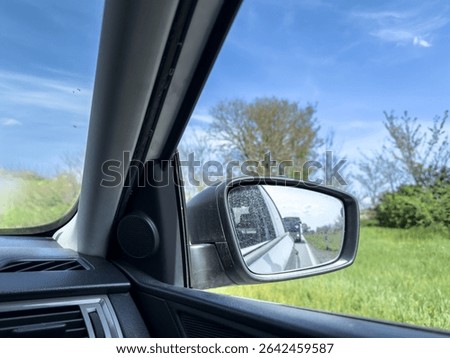Driver side view captures a dusty car mirror reflecting a bus approaching from behind on a rural two lane road, framed by green and spring trees under a clear blue sky, conveying road safety awareness
