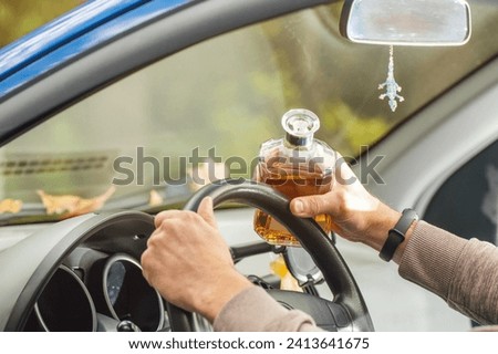 The driver is sitting behind the wheel and holding a bottle of alcoholic drink. Breaking the law and drinking alcohol while driving.