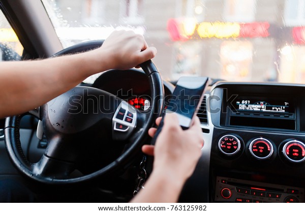 Driver with a smartphone in hand in the driver\'s\
seat. The concept of inattention at the wheel. Driving with one\
hand, distracted by SMS or call by mobile. Cropped hand of\
businessman using phone