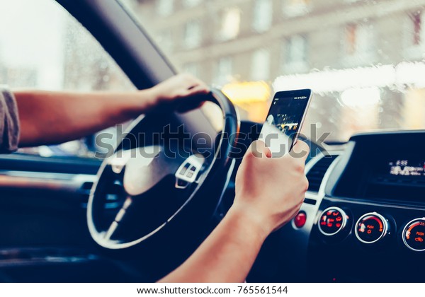 Driver with a smartphone in hand in the driver's
seat. The concept of inattention at the wheel. Distracted by SMS or
call by mobile. Cropped hand of businessman using phone. Vintage
style photo