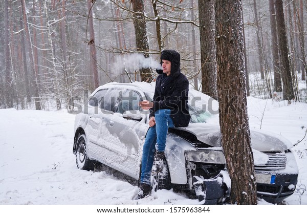 The driver smokes a cigarette next to the\
wrecked car. Accident in the snowy\
forest