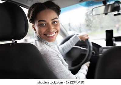 Driver turns to passenger seat and smiles at camera. Happy millennial african american woman taxi driver sitting in car holding to steering wheel, next to smartphone with blank screen, panorama