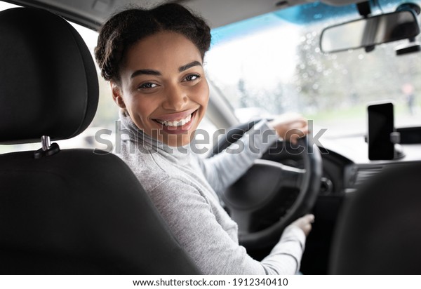 Driver turns to passenger seat and smiles at\
camera. Happy millennial african american woman taxi driver sitting\
in car holding to steering wheel, next to smartphone with blank\
screen, panorama