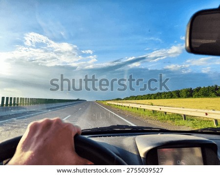A driver views the empty road ahead stretching into the countryside. Green fields and blue skies fill the scene during the road trip.